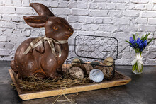 Brown Clay Easter Bunny In Wooden Bowl With Easter Eggs And Hay In Front Of White Brick Wall