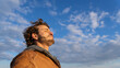 Young man taking a deep breath. He is outdoors with blue sky and clouds in the background. He takes a breath facing the sun with his eyes closed and the wind in his face. Copy space on the right