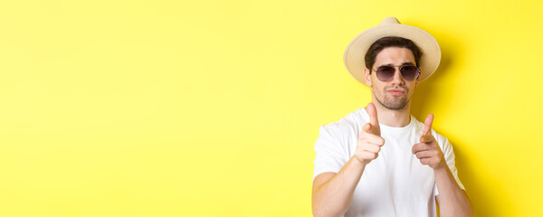 Wall Mural - Concept of tourism and vacation. Close-up of cool guy in summer hat and sunglasses pointing finger guns at camera, standing over yellow background