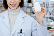 © InfiniteFlow - Closeup young affable pharmacist show qualified medicine pill bottle mockup product for copyspace at pharmacy. Druggist working with her diary job at drugstore, medicine box on shelf at background