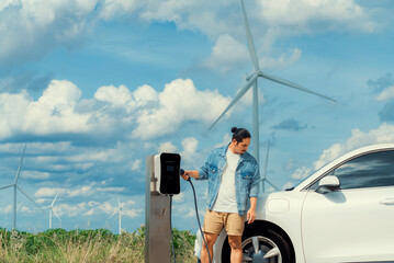 Progressive man with his electric car, EV car recharging energy from charging station on green field with wind turbine as concept of future sustainable energy. Electric vehicle with energy generator.