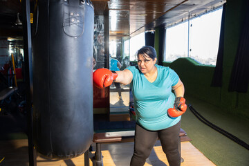 Wall Mural - Young overweight indian woman wearing boxing gloves punching bag in gym, Plus size sporty asian female workout to lose weight. Fitness and healthcare.