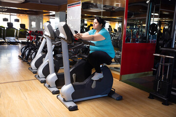 Wall Mural - Young overweight indian woman is workout in gym to lose weight. Asian female doing cardio training on bicycle machine. Full length.