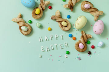 Wall Mural - Colors Easter eggs with Easter rabbit-shaped buns puff pastry with cinnamon on pastel green background. Happy Easter Holidays. View from above.