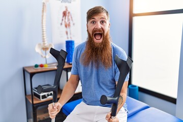 Wall Mural - Redhead man with long beard holding crutches at rehabilitation clinic celebrating crazy and amazed for success with open eyes screaming excited.