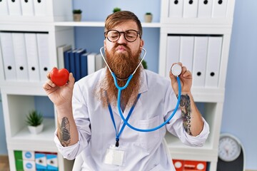 Wall Mural - Redhead man with long beard wearing doctor uniform holding heart and stethoscope looking at the camera blowing a kiss being lovely and sexy. love expression.