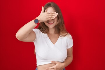 Sticker - Young caucasian woman standing over red background smiling and laughing with hand on face covering eyes for surprise. blind concept.