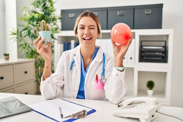 Poster - Young blonde woman wearing doctor uniform holding balloon and cactus winking looking at the camera with sexy expression, cheerful and happy face.