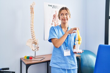 Poster - Young blonde orthopedic woman holding anatomical model of knee joint smiling looking to the side and staring away thinking.