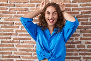 Poster - Beautiful brunette woman standing over bricks wall posing funny and crazy with fingers on head as bunny ears, smiling cheerful