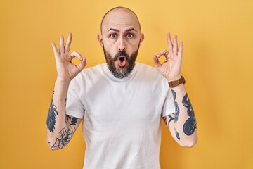 Poster - Young hispanic man with tattoos standing over yellow background looking surprised and shocked doing ok approval symbol with fingers. crazy expression