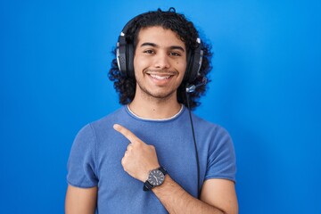 Hispanic man with curly hair listening to music using headphones cheerful with a smile of face pointing with hand and finger up to the side with happy and natural expression on face