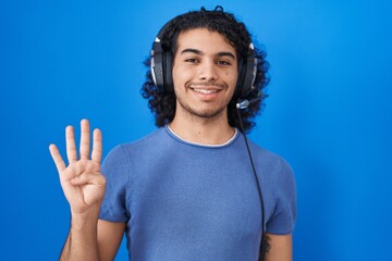 Wall Mural - Hispanic man with curly hair listening to music using headphones showing and pointing up with fingers number four while smiling confident and happy.