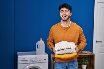 Wall Mural - Hispanic man with beard holding clean folded laundry smiling and laughing hard out loud because funny crazy joke.