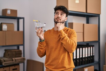 Sticker - Hispanic man with beard standing by manikin at small business holding cart serious face thinking about question with hand on chin, thoughtful about confusing idea