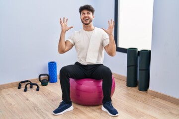 Wall Mural - Hispanic man with beard sitting on pilate balls at yoga room crazy and mad shouting and yelling with aggressive expression and arms raised. frustration concept.