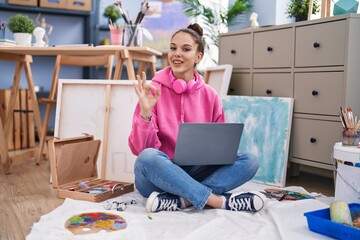 Canvas Print - Young hispanic girl using laptop at painter studio doing ok sign with fingers, smiling friendly gesturing excellent symbol