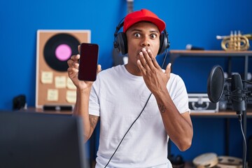 Wall Mural - Young hispanic man listening to music wearing headphones showing smartphone screen covering mouth with hand, shocked and afraid for mistake. surprised expression