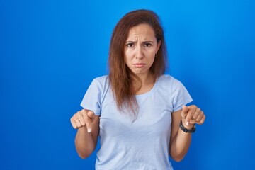 Sticker - Brunette woman standing over blue background pointing down looking sad and upset, indicating direction with fingers, unhappy and depressed.