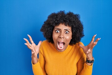 Sticker - Black woman with curly hair standing over blue background crazy and mad shouting and yelling with aggressive expression and arms raised. frustration concept.