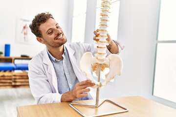 Canvas Print - Young hispanic man wearing physiotherapist uniform touching anatomical model of vertebral column at clinic