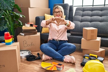 Sticker - Young hispanic woman moving to a new home sitting on the floor smiling in love doing heart symbol shape with hands. romantic concept.