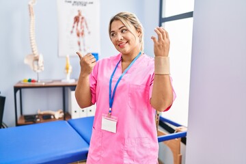 Sticker - Young hispanic woman working at pain recovery clinic wearing wristband pointing thumb up to the side smiling happy with open mouth