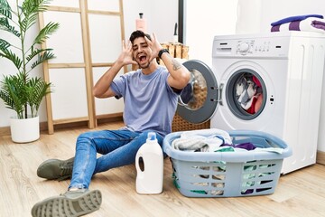 Wall Mural - Young hispanic man putting dirty laundry into washing machine smiling cheerful playing peek a boo with hands showing face. surprised and exited