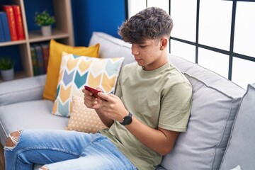 Poster - Young hispanic teenager watching video on smartphone sitting on sofa at home