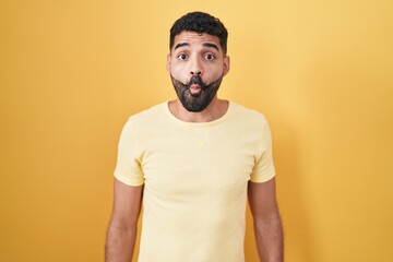 Sticker - Hispanic man with beard standing over yellow background making fish face with lips, crazy and comical gesture. funny expression.