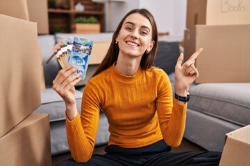 Poster - Young caucasian woman sitting on the floor at new home holding canadian money smiling happy pointing with hand and finger to the side