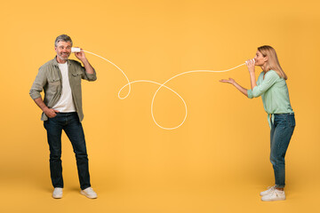 Communication and fun. Happy middle aged spouses talking through tin can telephone, yellow studio background