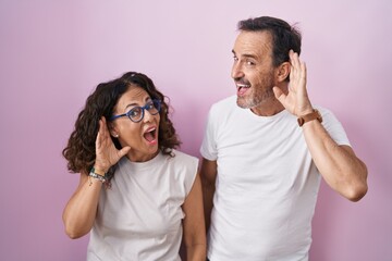 Poster - Middle age hispanic couple together over pink background smiling with hand over ear listening an hearing to rumor or gossip. deafness concept.