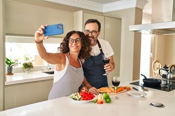 Wall Mural - Middle age hispanic couple smiling confident cooking and make selfie by the smartphone at kitchen