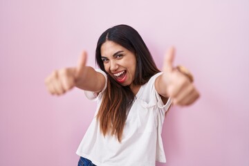 Wall Mural - Young arab woman standing over pink background approving doing positive gesture with hand, thumbs up smiling and happy for success. winner gesture.