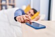 Young hispanic man sleeping on bed turning off smartphone alarm at bedroom