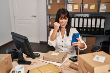 Wall Mural - Young brunette woman working at small business ecommerce using smartphone annoyed and frustrated shouting with anger, yelling crazy with anger and hand raised