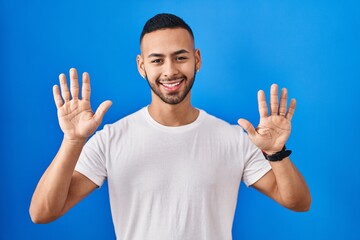 Poster - Young hispanic man standing over blue background showing and pointing up with fingers number ten while smiling confident and happy.