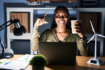 Canvas Print - African woman working using computer laptop at night smiling and confident gesturing with hand doing small size sign with fingers looking and the camera. measure concept.