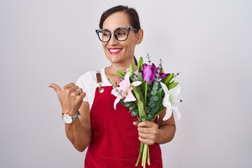 Poster - Middle age brunette woman wearing apron working at florist shop holding bouquet smiling with happy face looking and pointing to the side with thumb up.