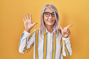 Poster - Middle age woman with grey hair standing over yellow background wearing glasses showing and pointing up with fingers number seven while smiling confident and happy.