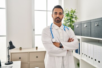 Wall Mural - Young hispanic man wearing doctor uniform standing with arms crossed gesture at clinic