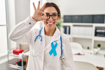 Wall Mural - Young brunette doctor woman wearing stethoscope at the clinic doing ok gesture with hand smiling, eye looking through fingers with happy face.