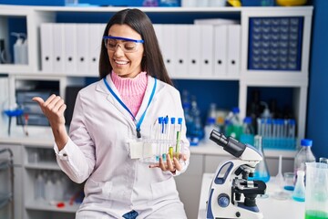 Poster - Young brunette woman working at scientist laboratory pointing thumb up to the side smiling happy with open mouth