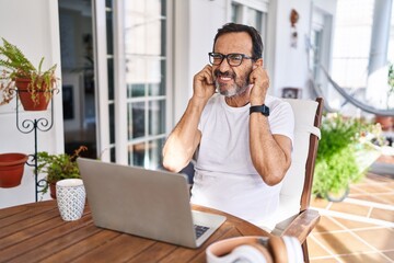 Wall Mural - Middle age man using computer laptop at home covering ears with fingers with annoyed expression for the noise of loud music. deaf concept.