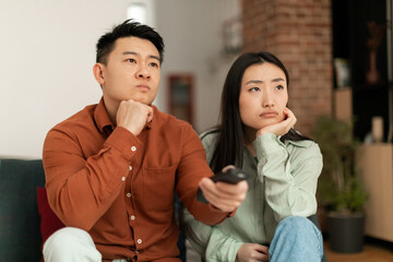 Wall Mural - Discontented asian couple watching television, pointing remote controller and switching TV channels, sitting on sofa