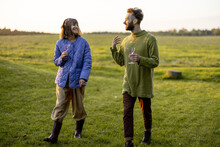 Stylish Man And Woman Dressed Warmly Have Conversation While Walk Together With Wine On Green Lawn During A Sunset. Couple Spend Leisure Autumn Time On Nature