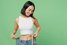 Young Fun Woman Wear White Clothes Show Loose Pants After Weightloss Hold Measure Tape On Waist Isolated On Plain Pastel Green Background. Proper Nutrition Healthy Fast Food Unhealthy Choice Concept.