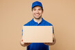 © ViDi Studio - Professional smiling positive fun delivery guy employee man wear blue cap t-shirt uniform workwear work as dealer courier hold cardboard box isolated on plain light beige background. Service concept.