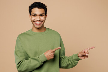 Wall Mural - Young man of African American ethnicity wears green sweatshirt point index finger aside indicate on workspace area copy space mock up isolated on plain pastel light beige background studio portrait.